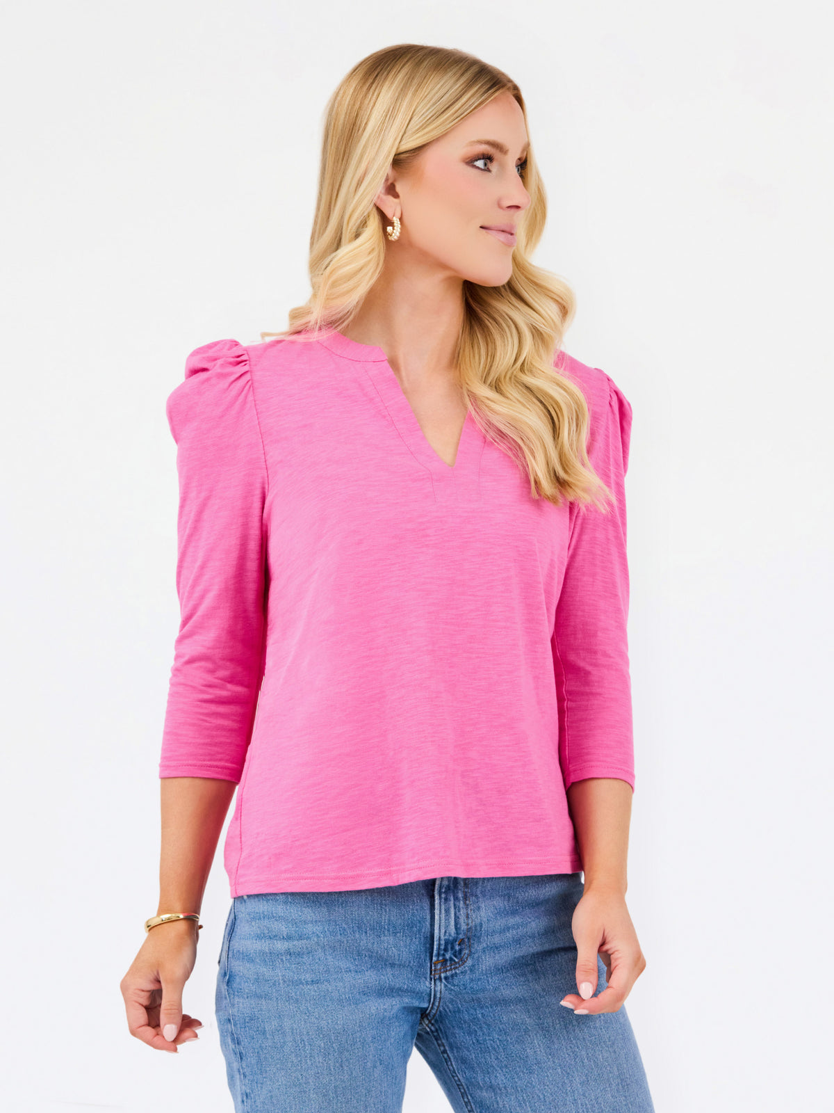 A blonde woman wearing a bright pink three-quarter sleeve top with puffed shoulders and a V-neckline, paired with blue jeans, photographed against a plain white studio background.