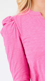 A close-up of a woman wearing a bright pink long-sleeve top with a gathered puff sleeve and a rounded neckline, shown against a plain white background.