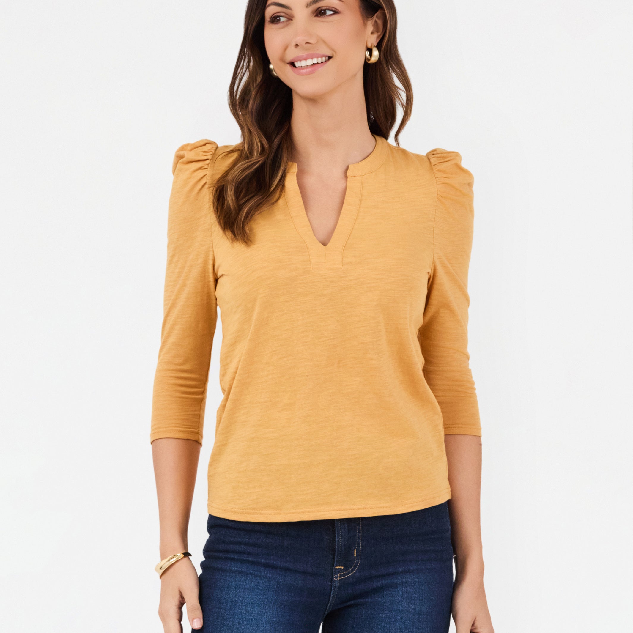 A woman modeling a mustard yellow three-quarter sleeve top with puffed shoulders and a split V-neckline, paired with dark blue jeans against a plain white background.
