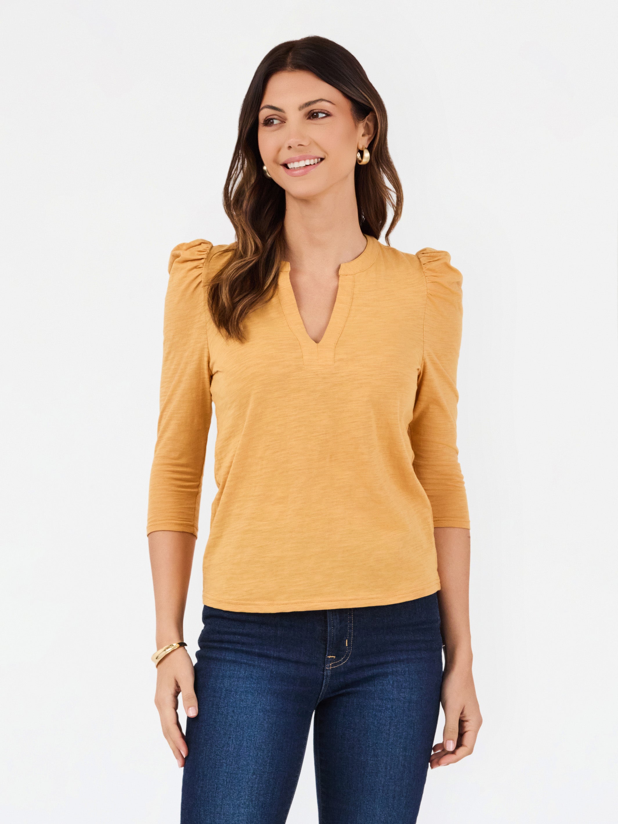 A woman modeling a mustard yellow three-quarter sleeve top with puffed shoulders and a split V-neckline, paired with dark blue jeans against a plain white background.
