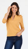 A woman modeling a mustard yellow knit top with three-quarter sleeves and puffed shoulders, paired with dark blue jeans, against a plain white studio background.