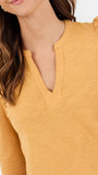 A close-up of a woman wearing a mustard yellow long-sleeve top with a split V-neckline and ruffled shoulders, shown against a plain light background.