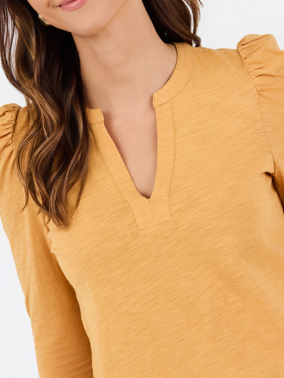A close-up of a woman wearing a mustard yellow long-sleeve top with a split V-neckline and ruffled shoulders, shown against a plain light background.