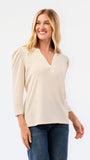 A blonde woman modeling a white long-sleeve knit top with a V-neck and puffed shoulders, paired with blue jeans, against a plain white studio background.
