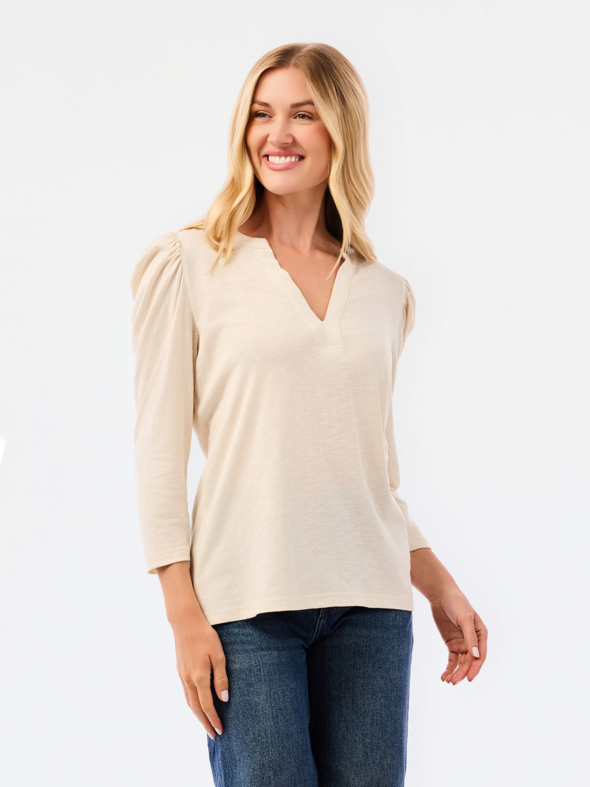 A blonde woman modeling a white long-sleeve knit top with a V-neck and puffed shoulders, paired with blue jeans, against a plain white studio background.