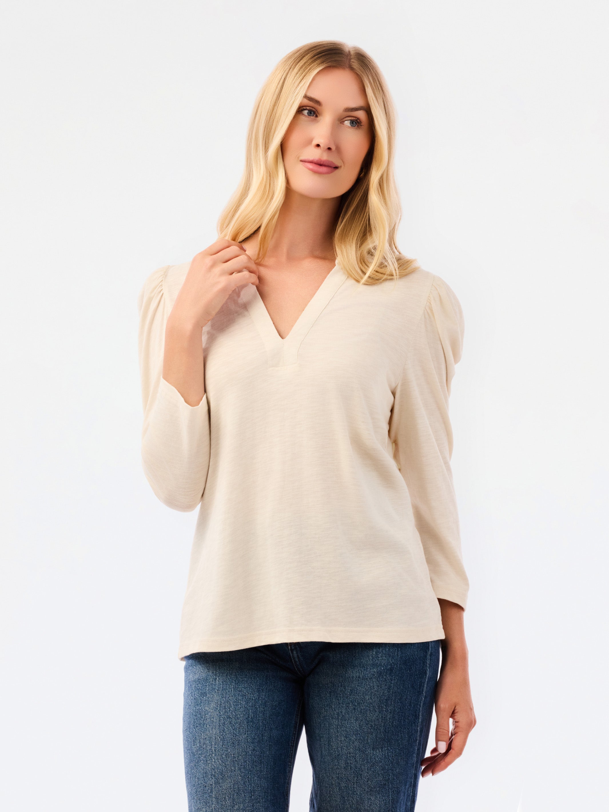A blonde woman modeling a white long-sleeve knit top with a V-neck and puffed shoulders, paired with blue jeans, against a plain white studio background.