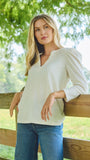 A woman with blonde hair wearing a light beige long-sleeve V-neck top and blue jeans, leaning against a wooden fence in an outdoor setting with green trees in the background.