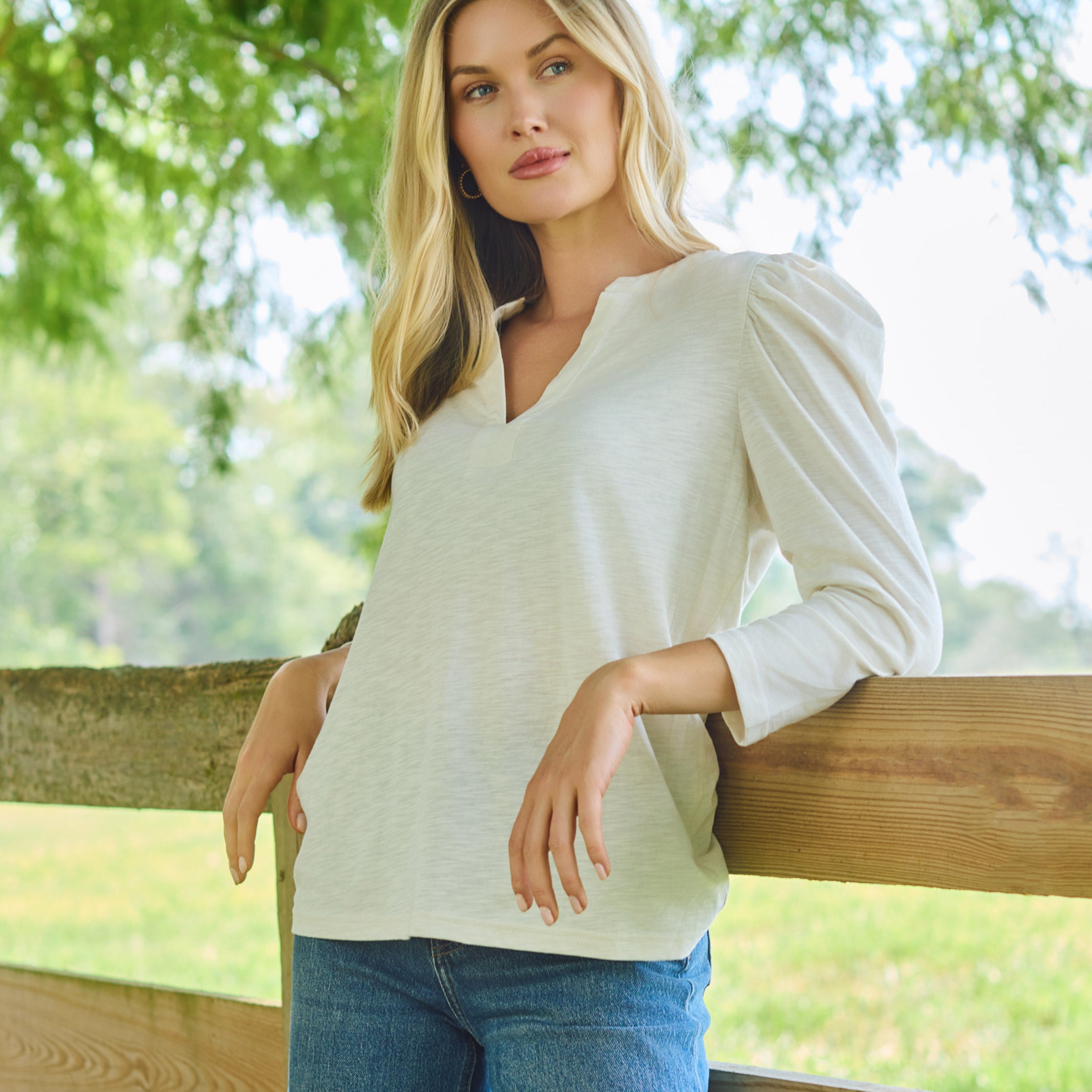 A woman with blonde hair wearing a light beige long-sleeve V-neck top and blue jeans, leaning against a wooden fence in an outdoor setting with green trees in the background.