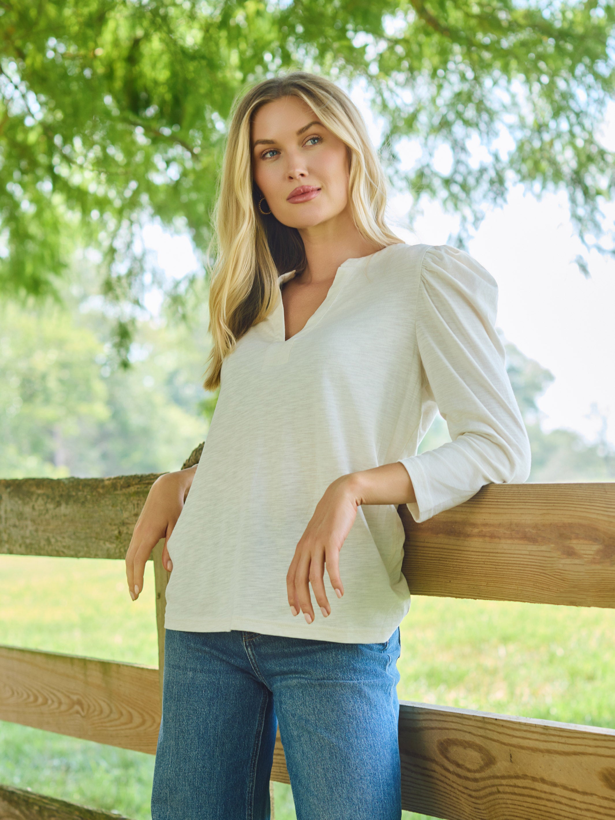 A woman with blonde hair wearing a light beige long-sleeve V-neck top and blue jeans, leaning against a wooden fence in an outdoor setting with green trees in the background.