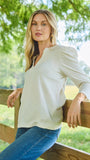 A woman wearing a light beige long-sleeve top with puffed shoulders and blue jeans, leaning against a wooden fence in an outdoor park setting with green trees in the background.