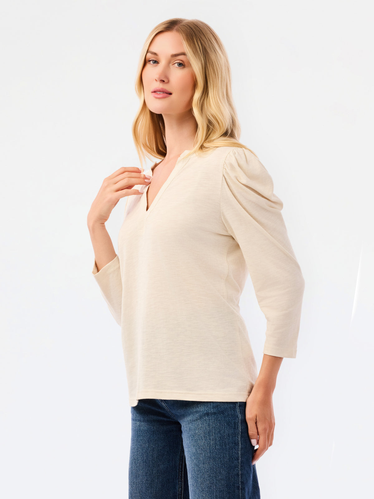 A blonde woman modeling a white long-sleeve knit top with a V-neck and puffed shoulders, paired with blue jeans, against a plain white studio background.