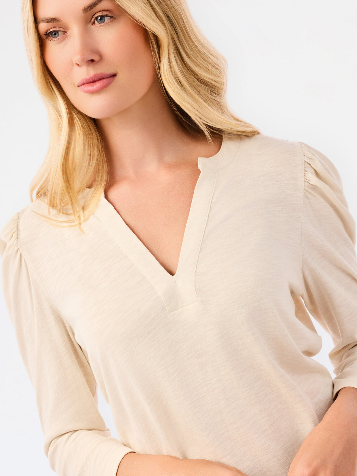 A blonde woman modeling a white long-sleeve knit top with a V-neck and puffed shoulders, paired with blue jeans, against a plain white studio background.