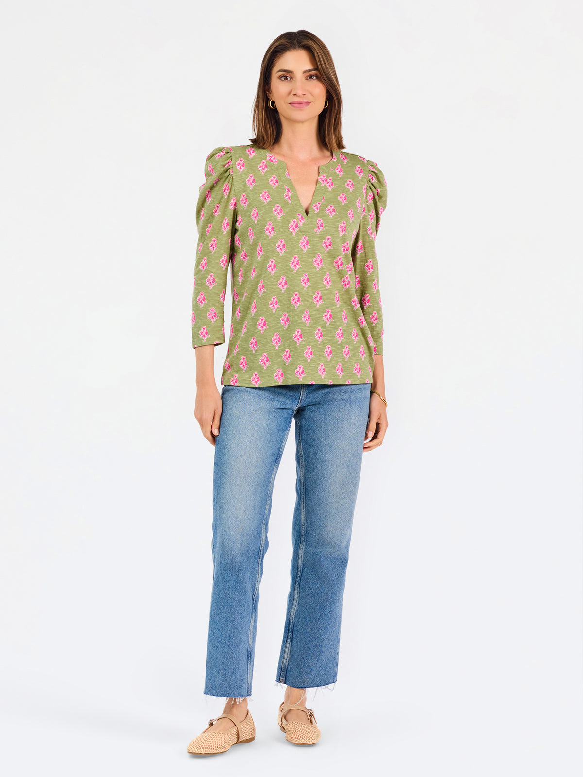 A woman modeling a green long-sleeve blouse with a pink floral pattern, puff sleeves, and a split V-neckline, paired with blue jeans against a plain white background.