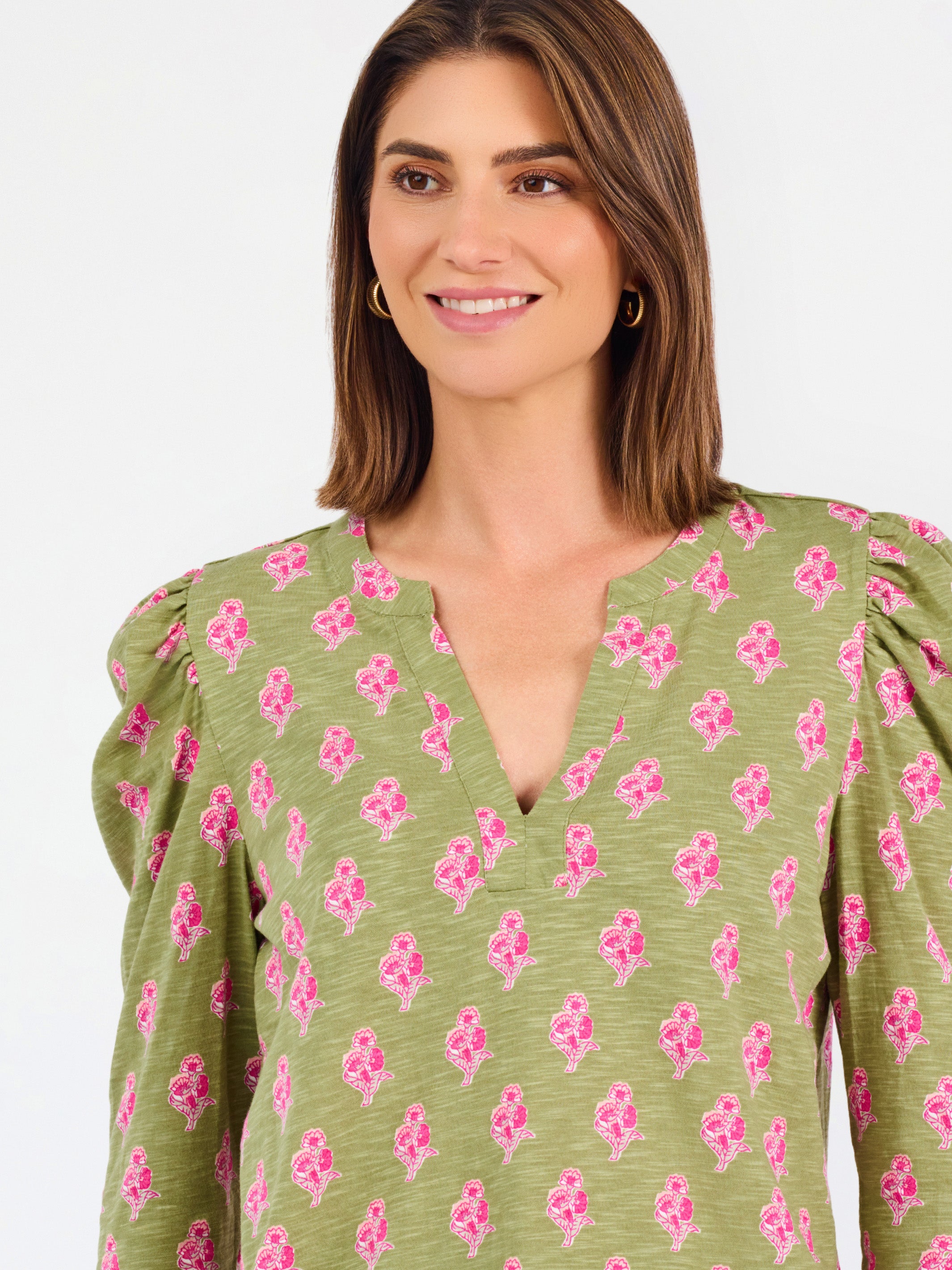 A woman modeling a green long-sleeve blouse with a pink floral pattern, puff sleeves, and a split V-neckline, paired with blue jeans against a plain white background.