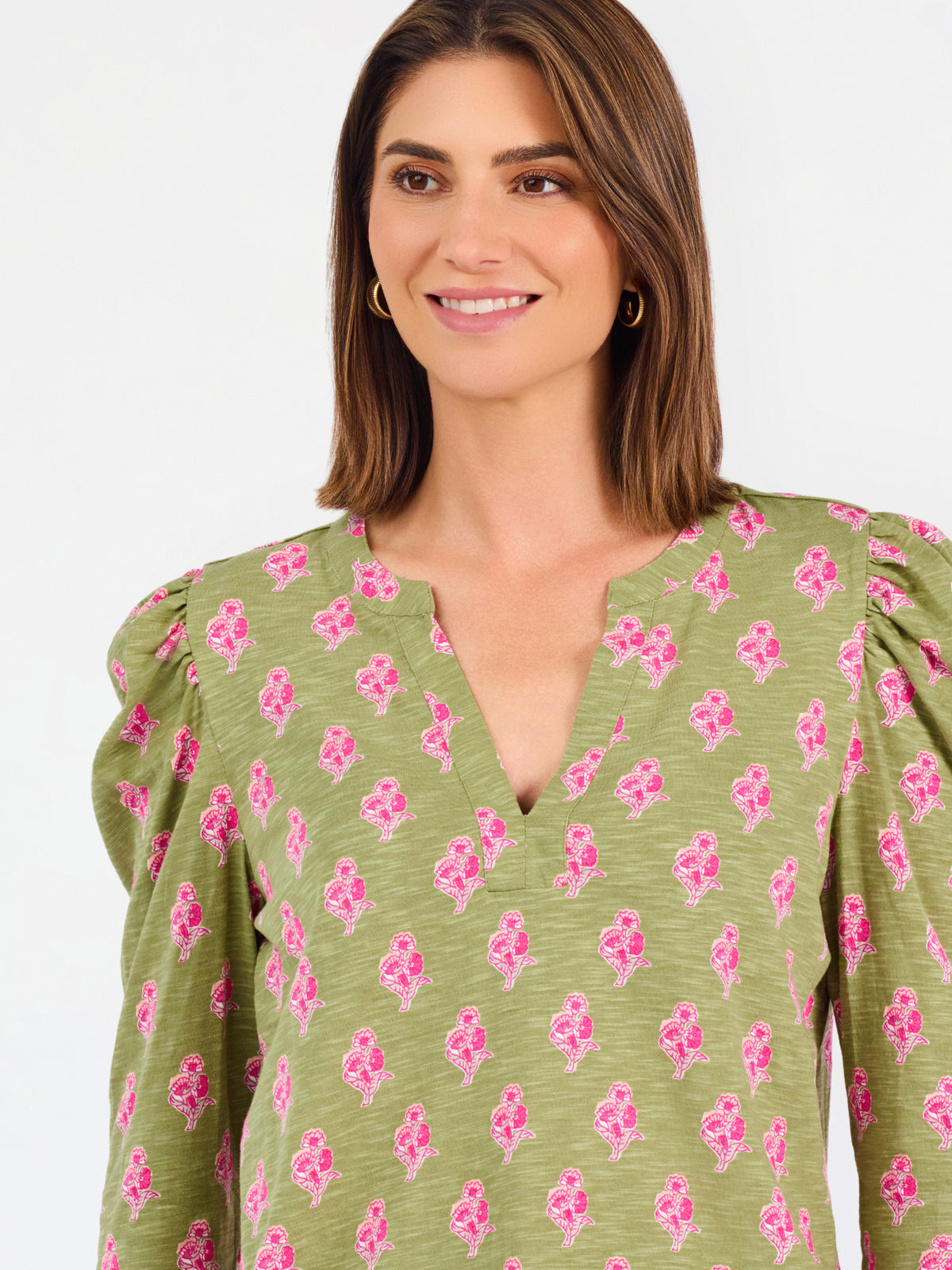 A woman modeling a green long-sleeve blouse with a pink floral pattern, puff sleeves, and a split V-neckline, paired with blue jeans against a plain white background.