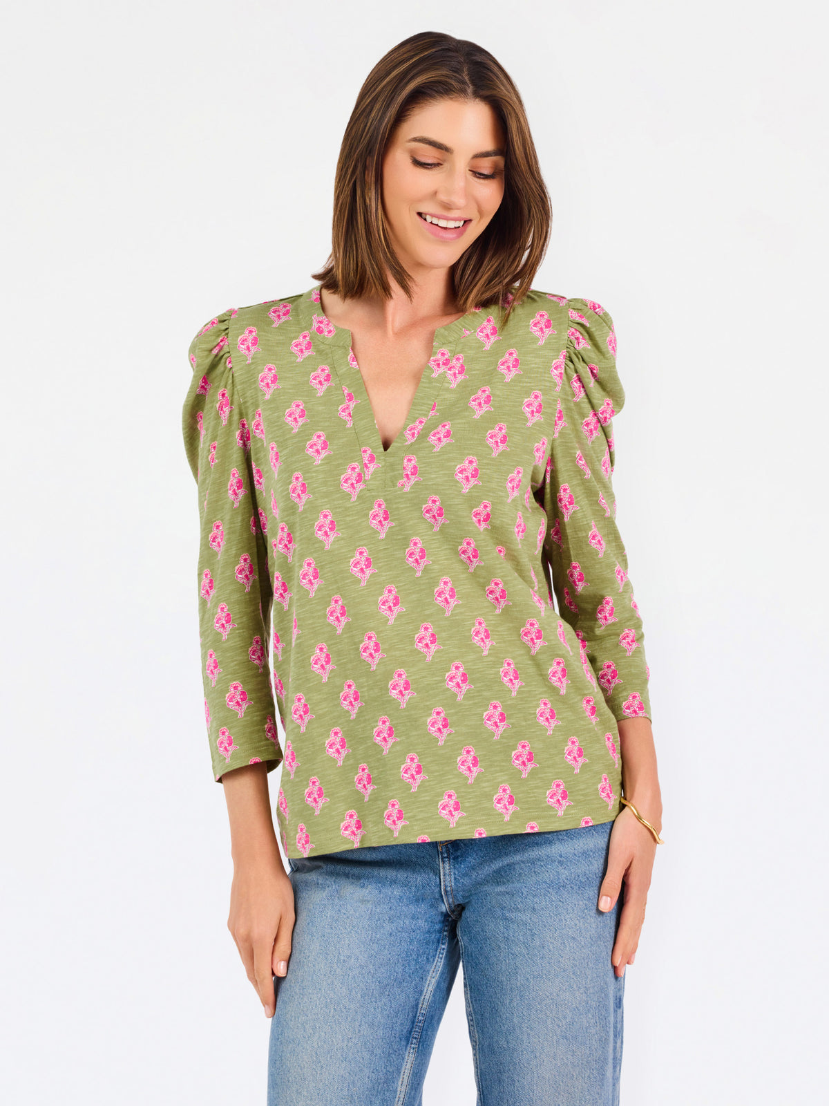 A woman modeling a green long-sleeve blouse with a pink floral pattern, puff sleeves, and a split V-neckline, paired with blue jeans against a plain white background.