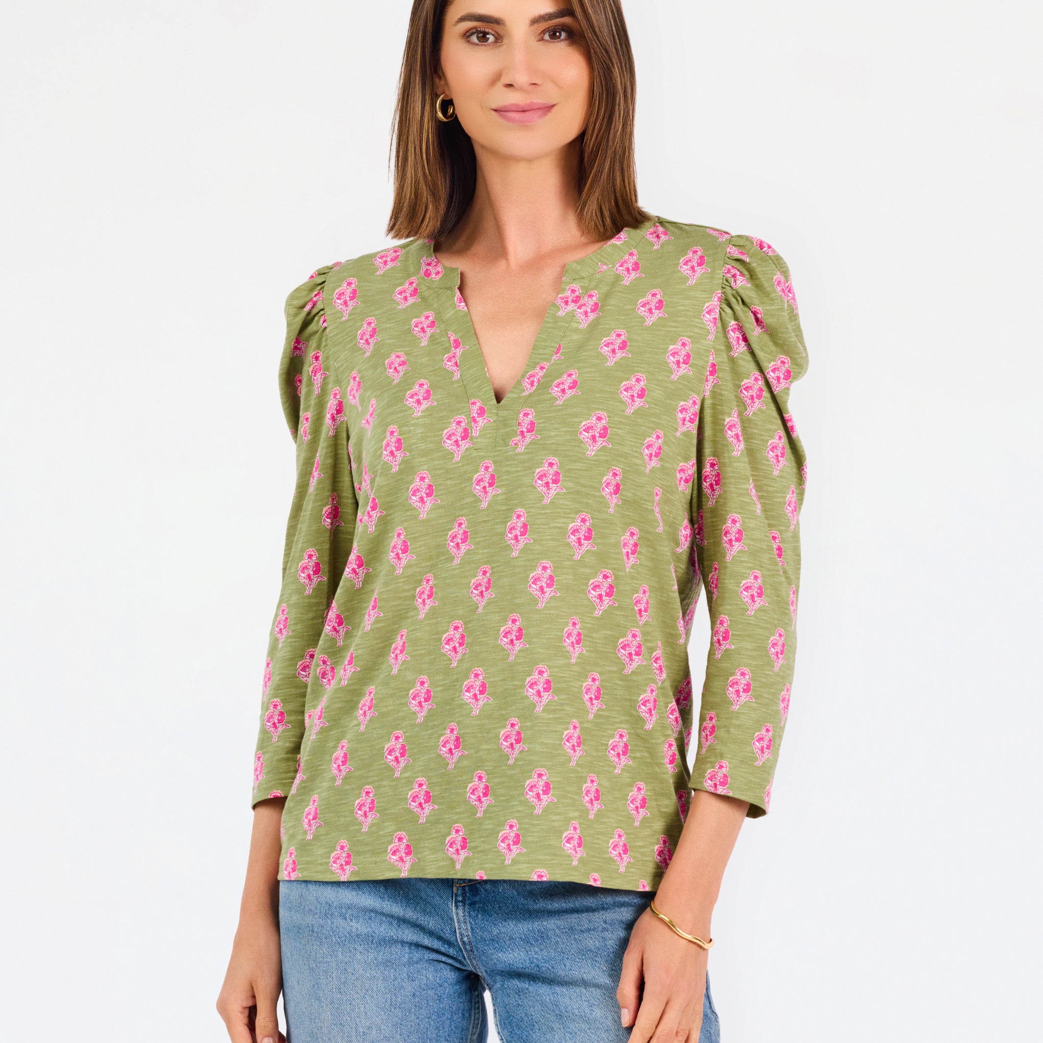 A woman modeling a green long-sleeve blouse with a pink floral pattern, puff sleeves, and a split V-neckline, paired with blue jeans against a plain white background.