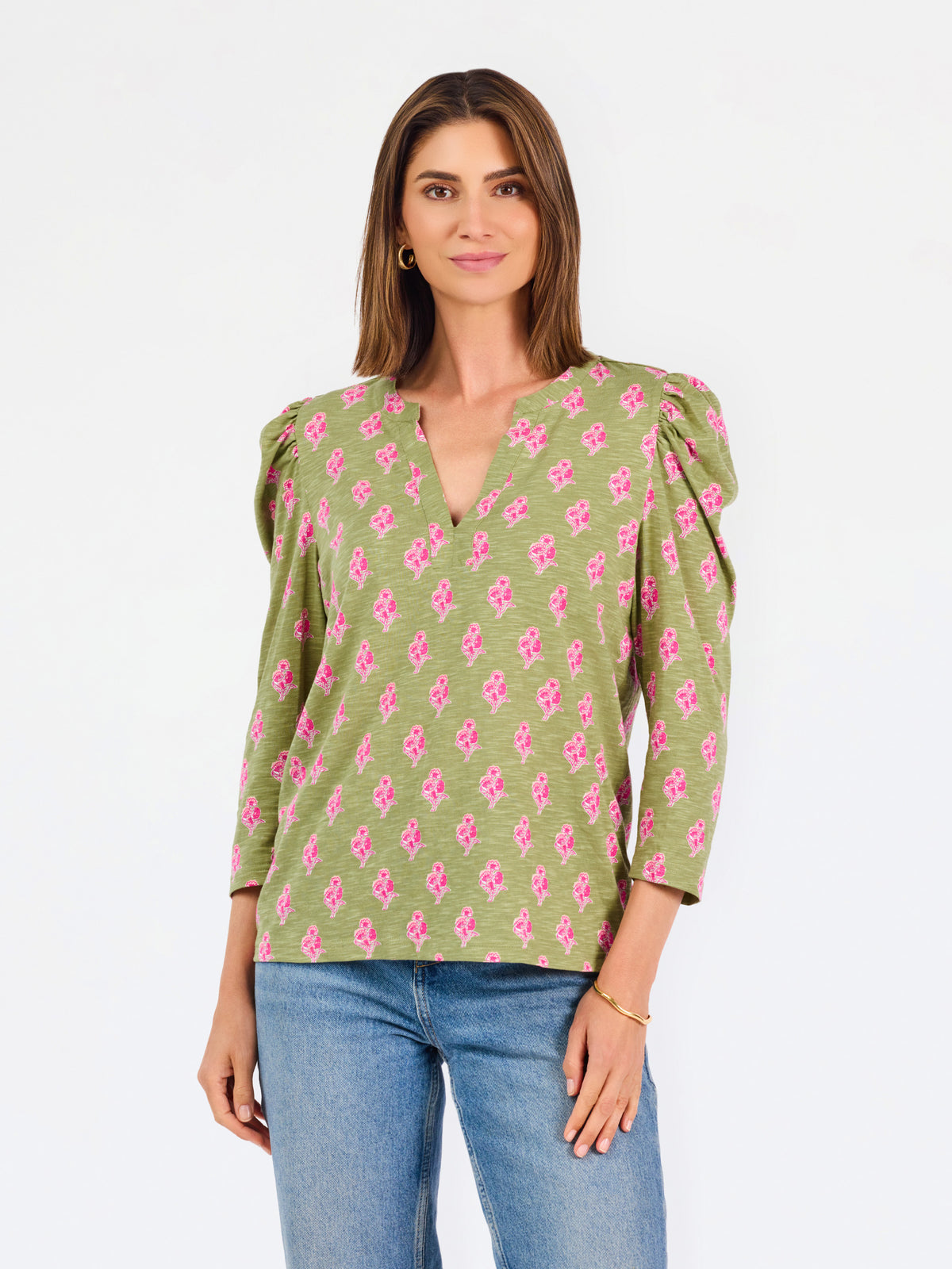 A woman modeling a green long-sleeve blouse with a pink floral pattern, puff sleeves, and a split V-neckline, paired with blue jeans against a plain white background.