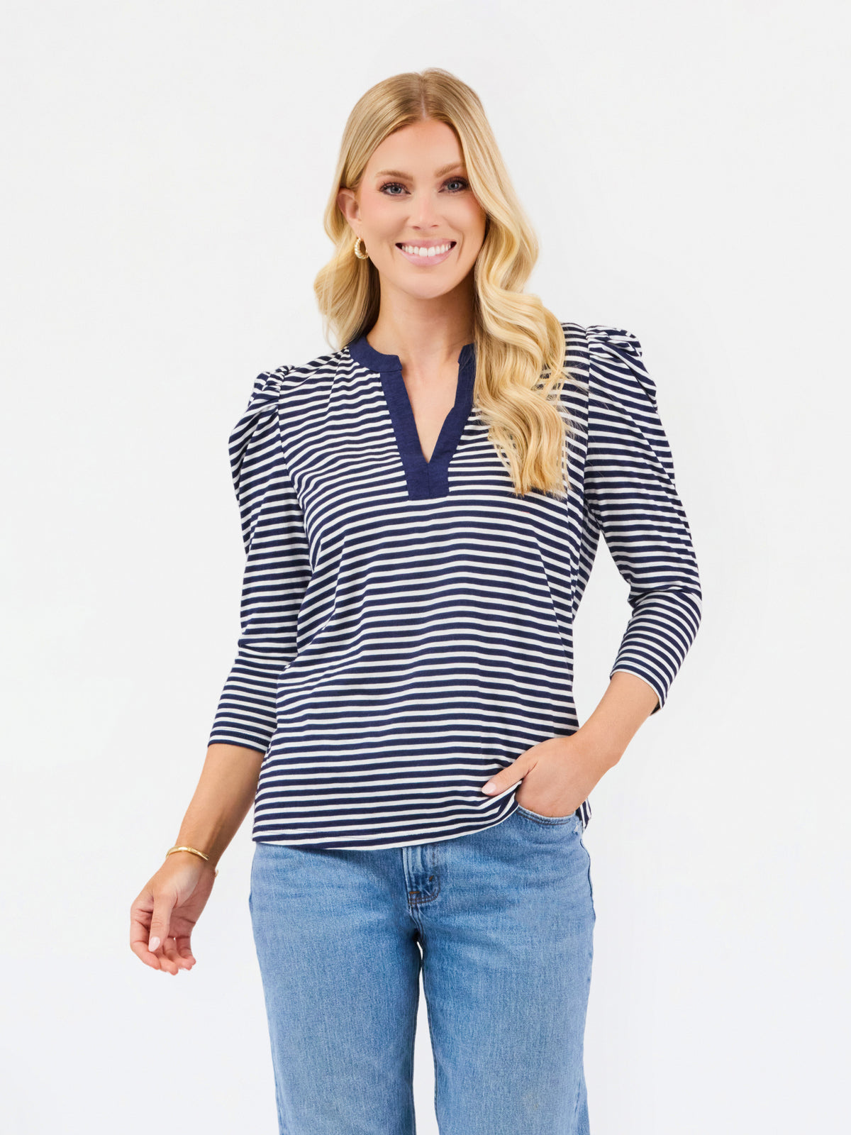 A woman modeling a navy and white horizontally striped long-sleeve top with a split V-neckline, paired with light blue jeans, against a plain white studio background.