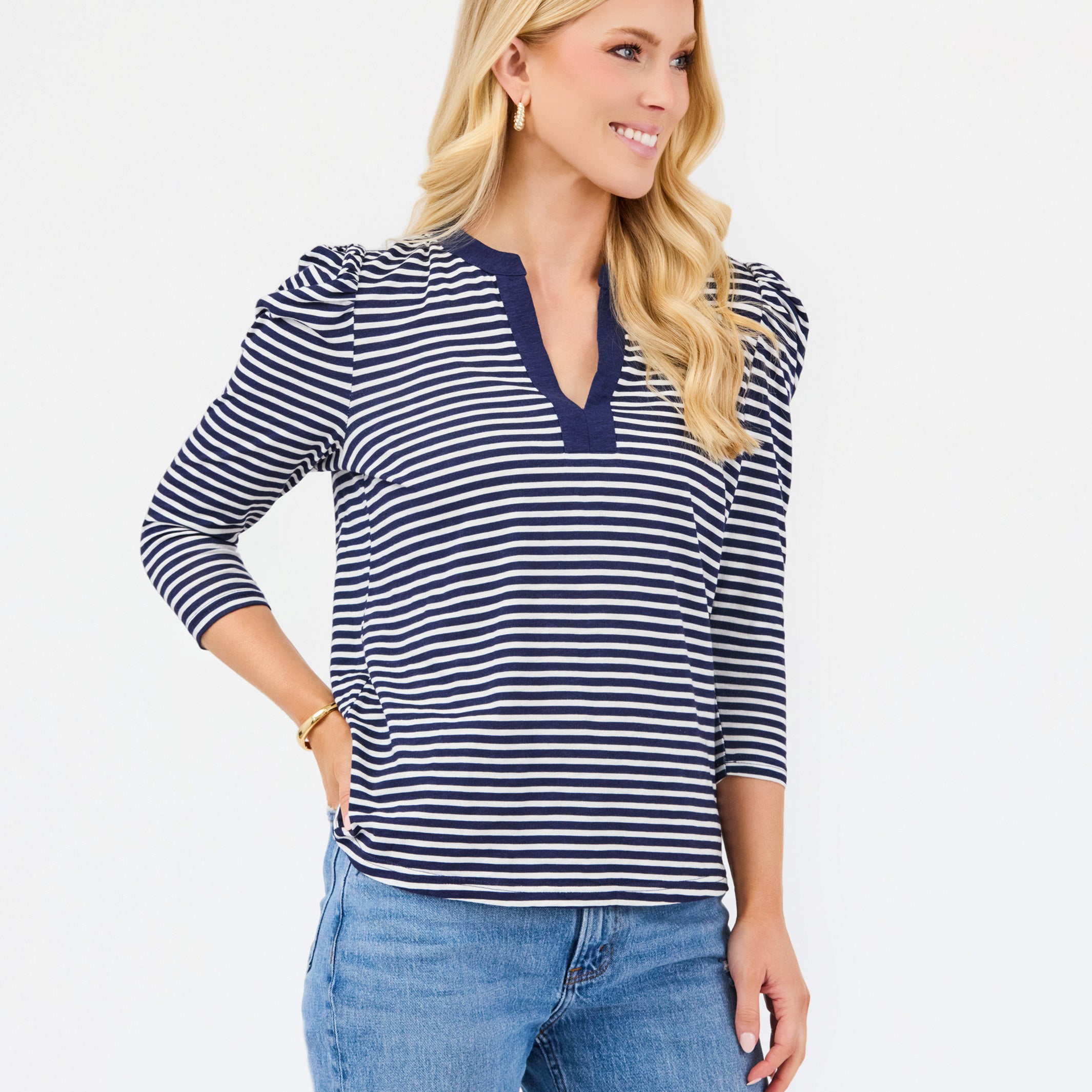 A blonde woman modeling a navy and white horizontally striped long-sleeve top with a split V-neckline, paired with blue jeans, against a plain white studio background.