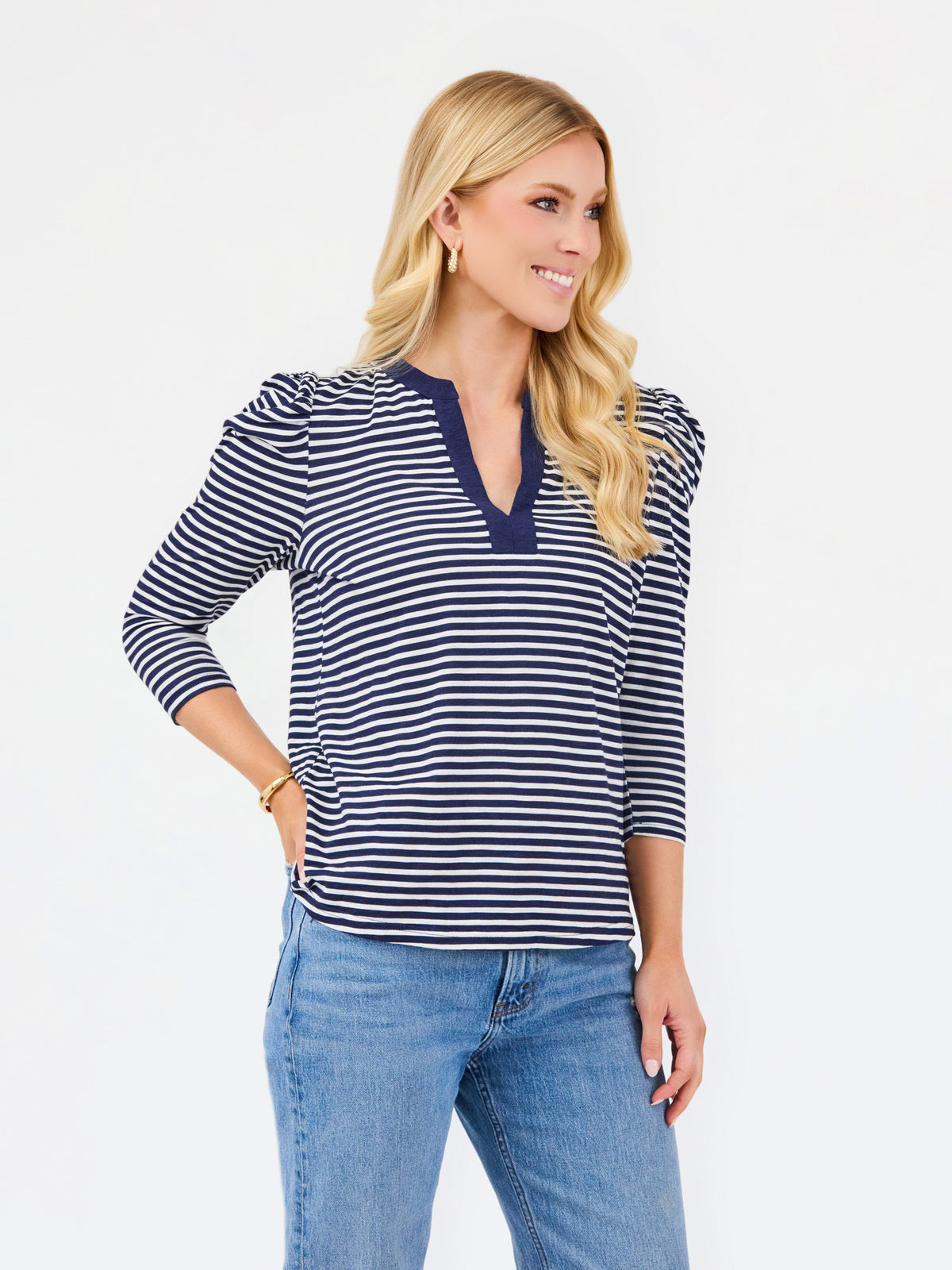 A blonde woman modeling a navy and white horizontally striped long-sleeve top with a split V-neckline, paired with blue jeans, against a plain white studio background.