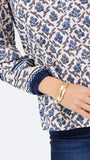 A close-up of a woman wearing a long-sleeve blouse with a blue and purple floral block print on a cream background, paired with dark blue jeans and a gold bangle.