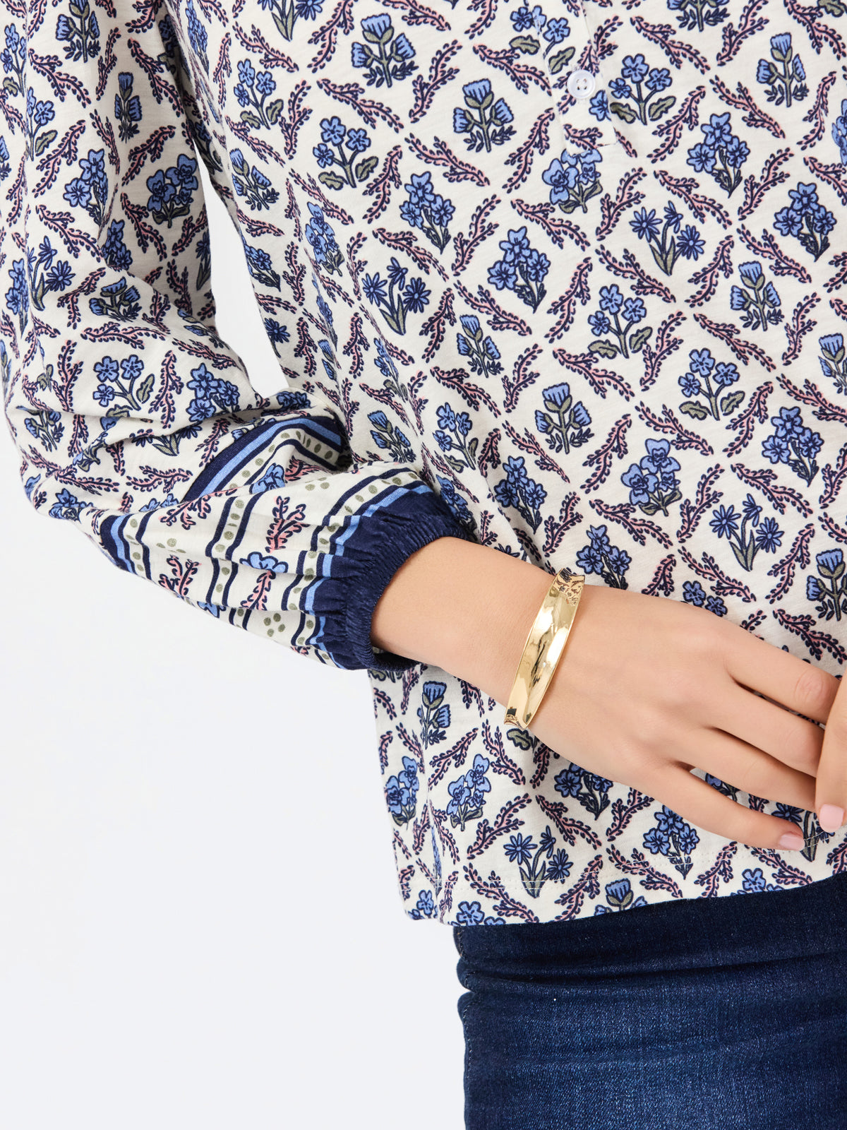 A close-up of a woman wearing a long-sleeve blouse with a blue and purple floral block print on a cream background, paired with dark blue jeans and a gold bangle.