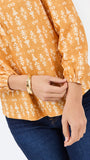 A close-up of a person wearing a mustard yellow long-sleeve blouse with a white floral block print pattern and dark blue jeans, accessorized with a gold bangle.