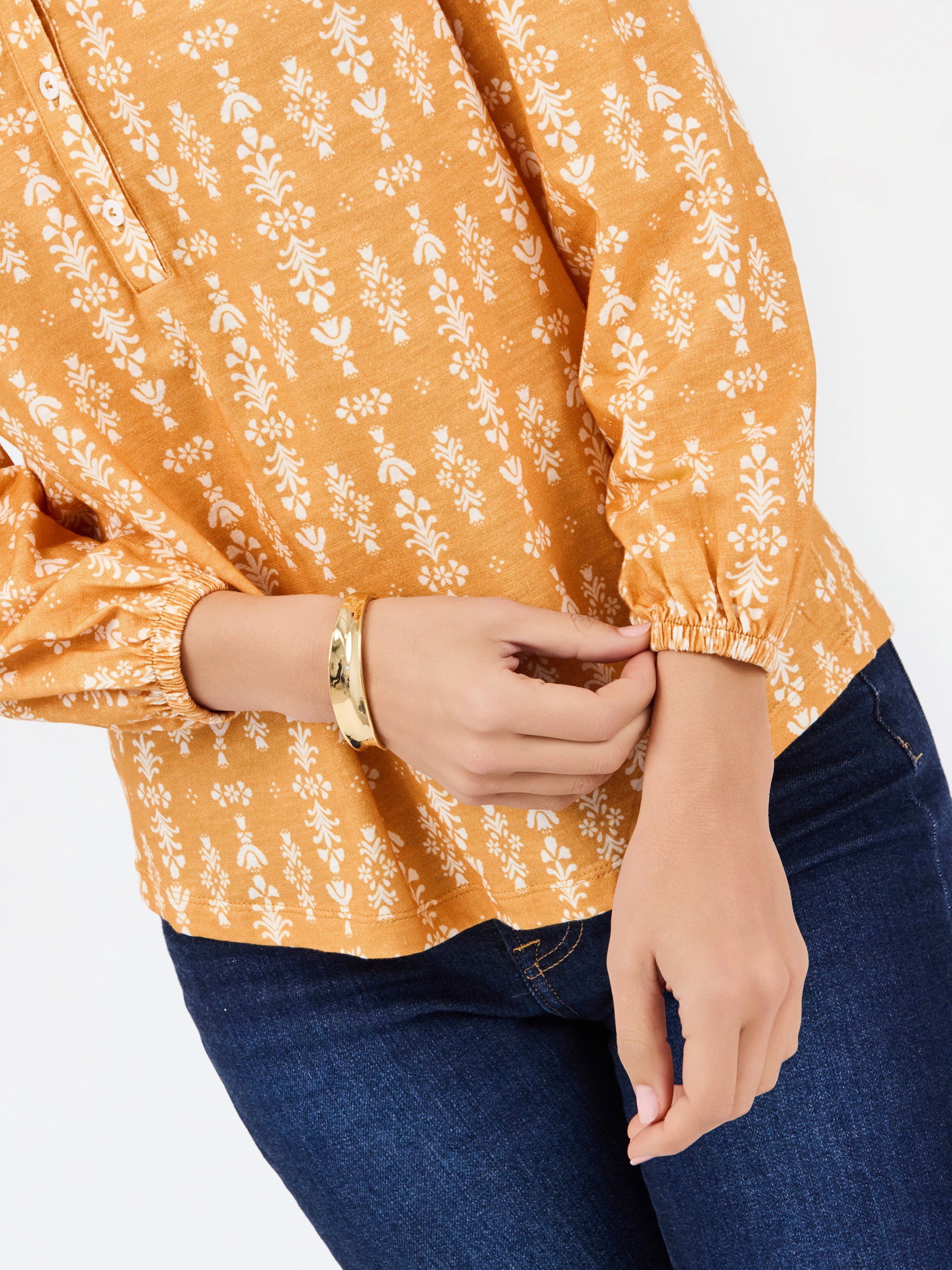 A close-up of a person wearing a mustard yellow long-sleeve blouse with a white floral block print pattern and dark blue jeans, accessorized with a gold bangle.