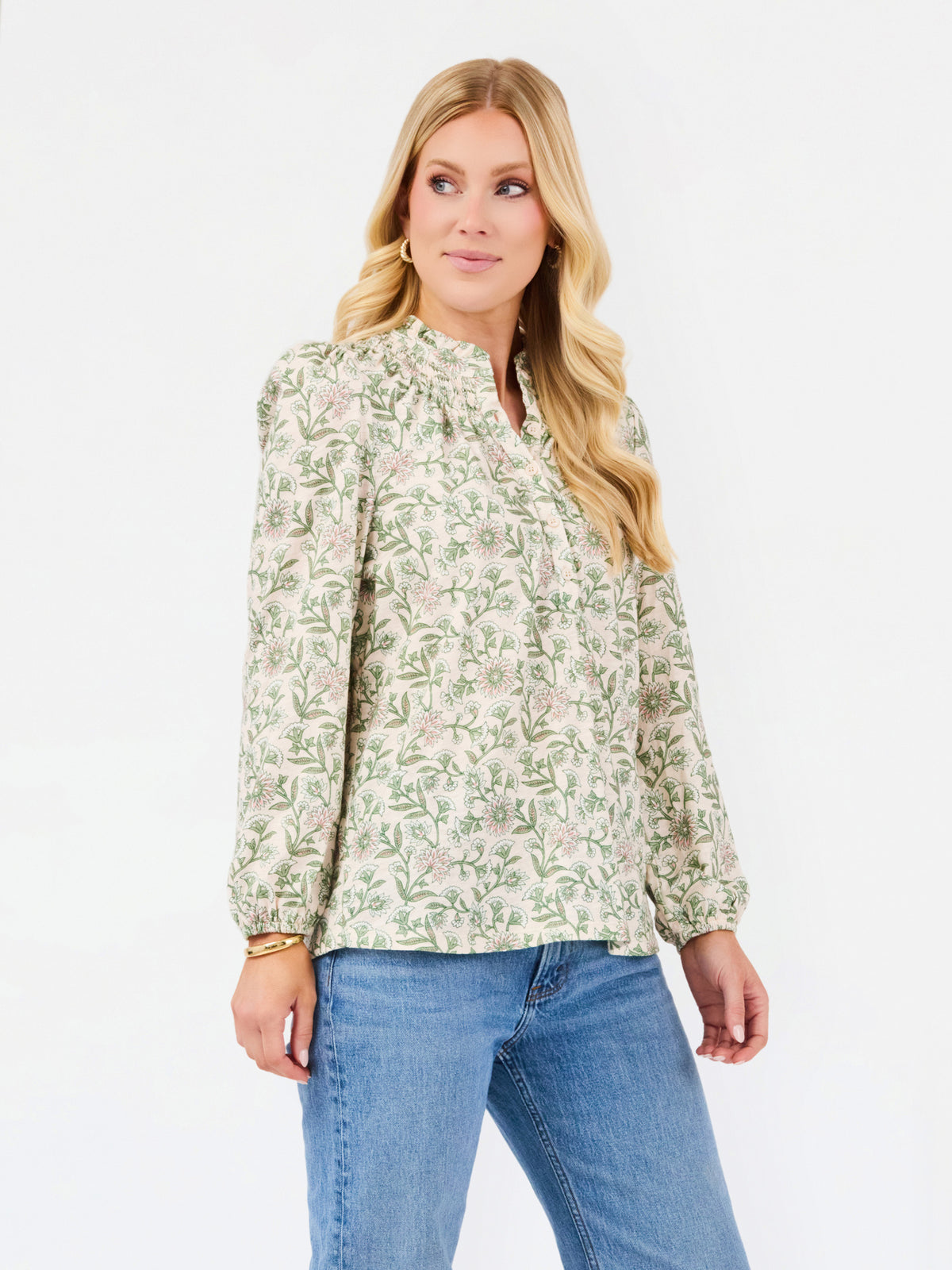 A woman modeling a long-sleeve blouse with a green and beige floral pattern, paired with blue jeans, against a plain white studio background.