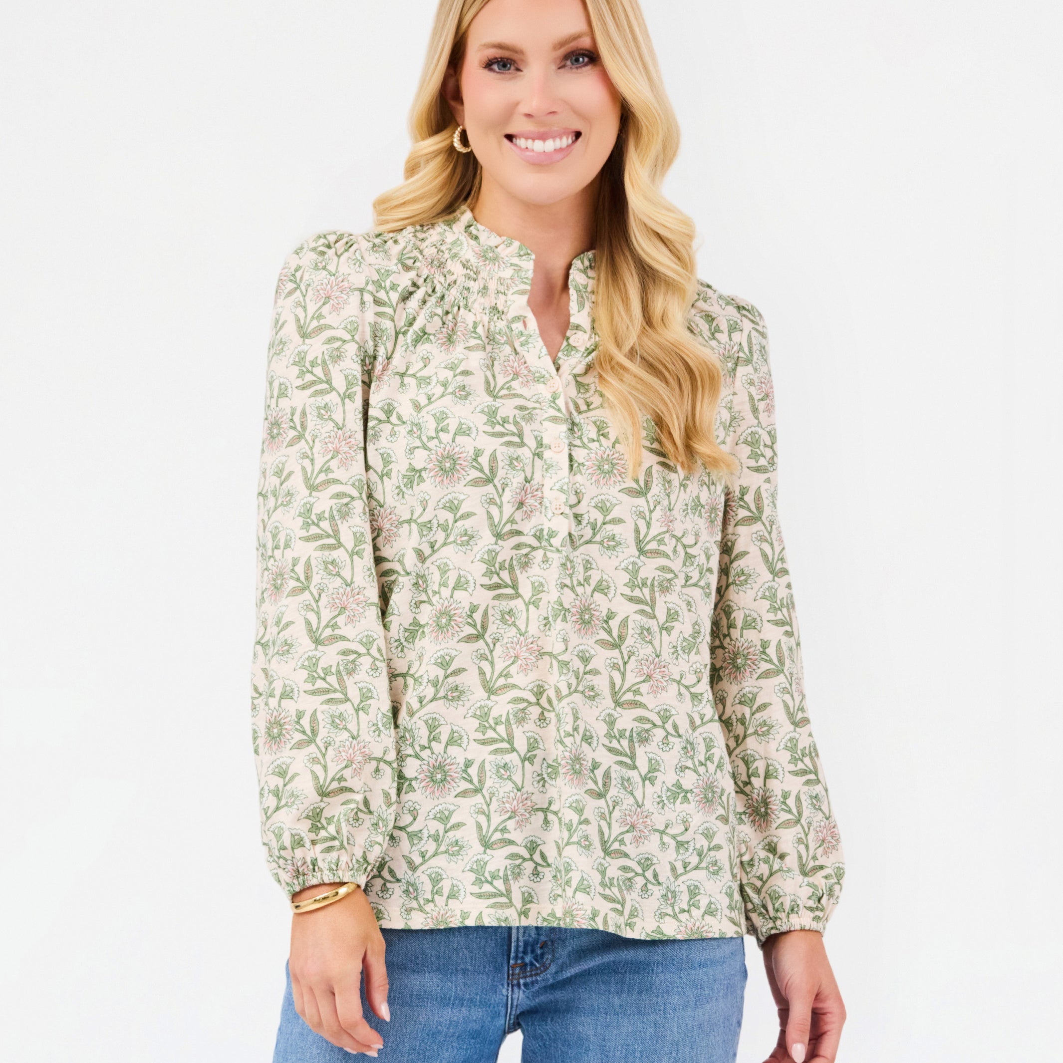 A woman modeling a long-sleeve blouse with a green and beige floral pattern, paired with blue jeans, against a plain white studio background.