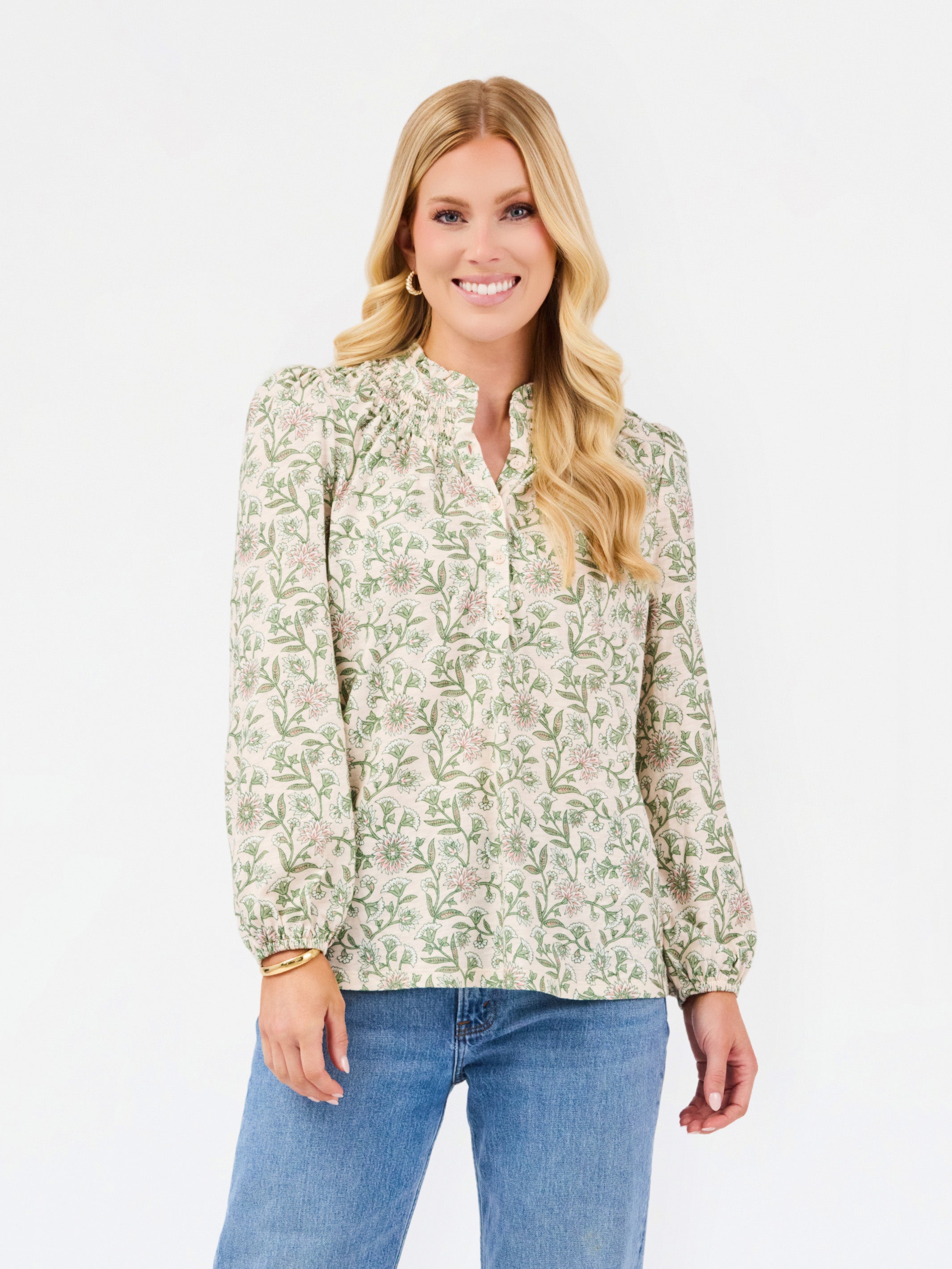 A woman modeling a long-sleeve blouse with a green and beige floral pattern, paired with blue jeans, against a plain white studio background.