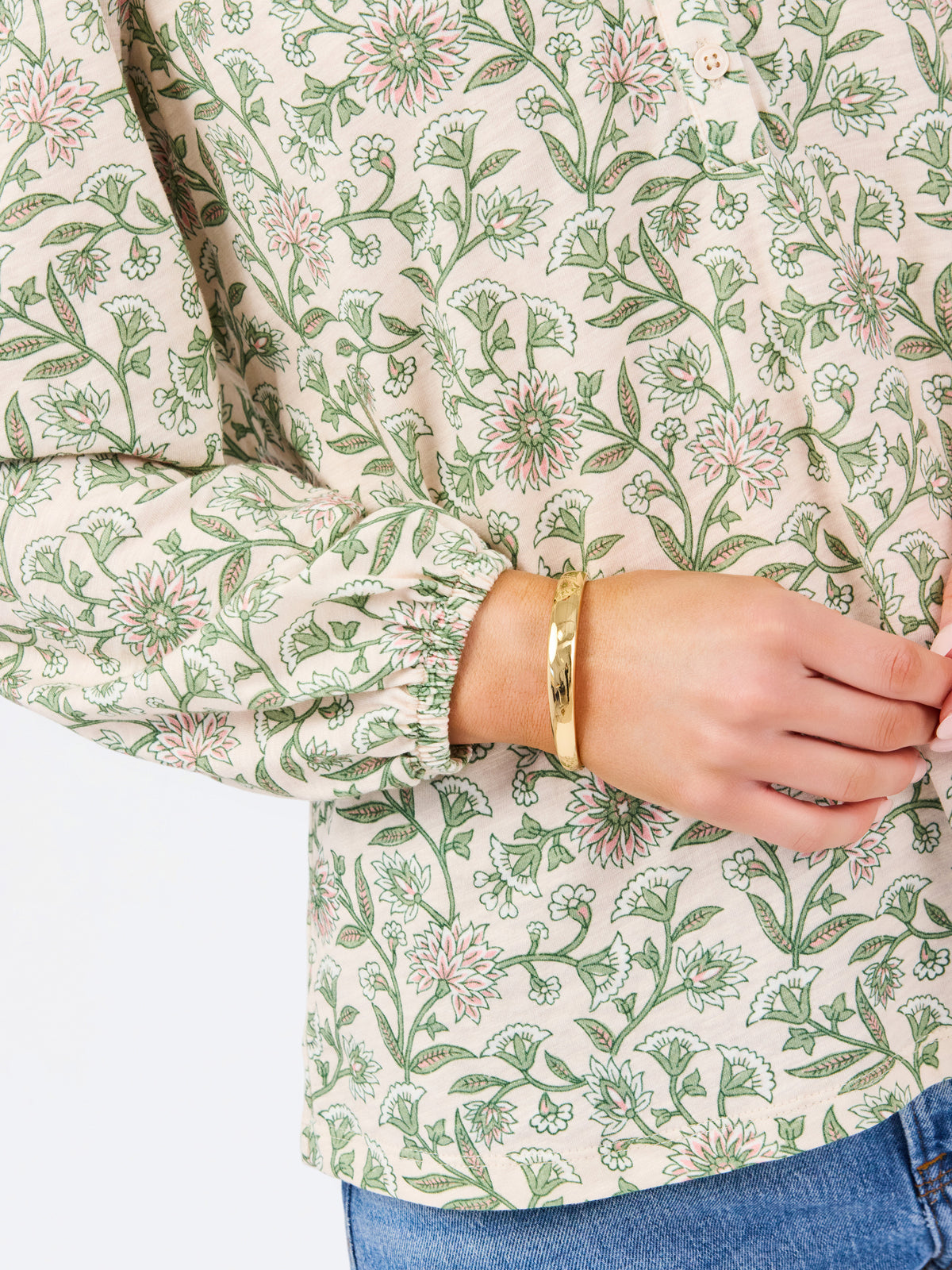 A woman modeling a long-sleeve blouse with a green and beige floral pattern, paired with blue jeans, against a plain white studio background.