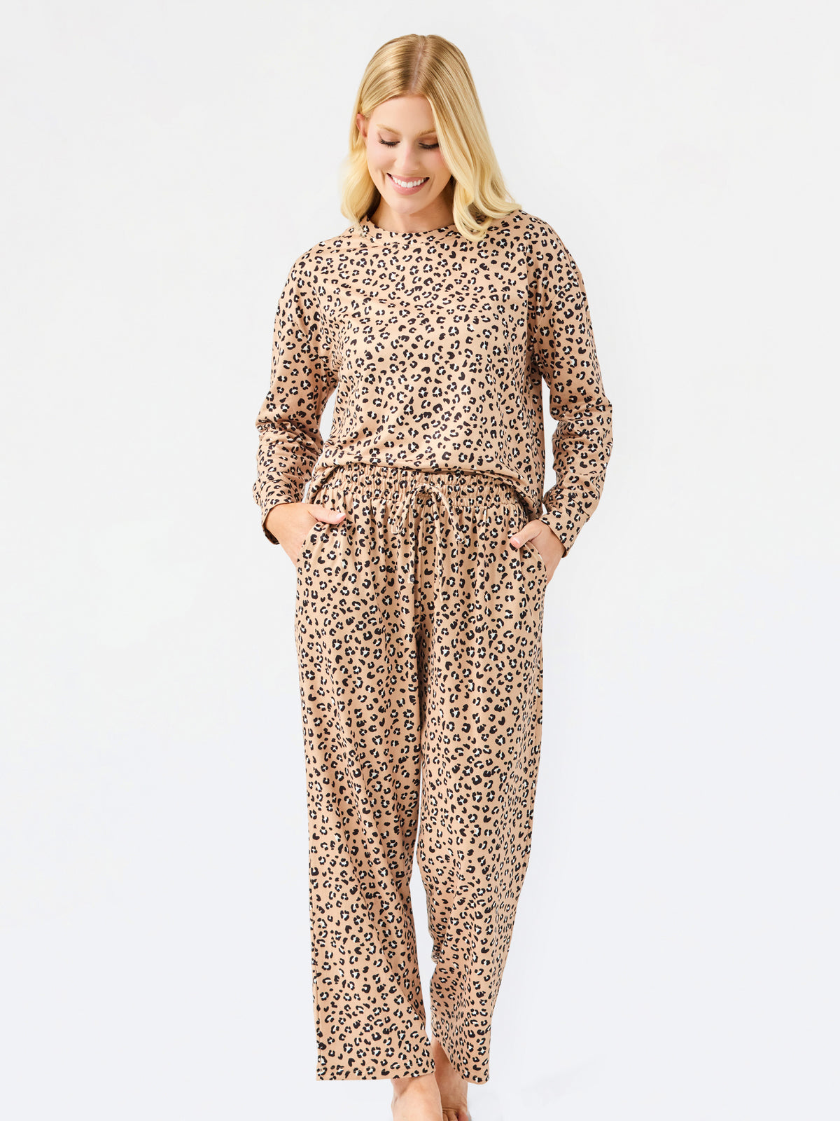 A woman modeling a matching two-piece leopard print pajama set with a long-sleeve top and wide-leg pants, standing against a plain white studio background.