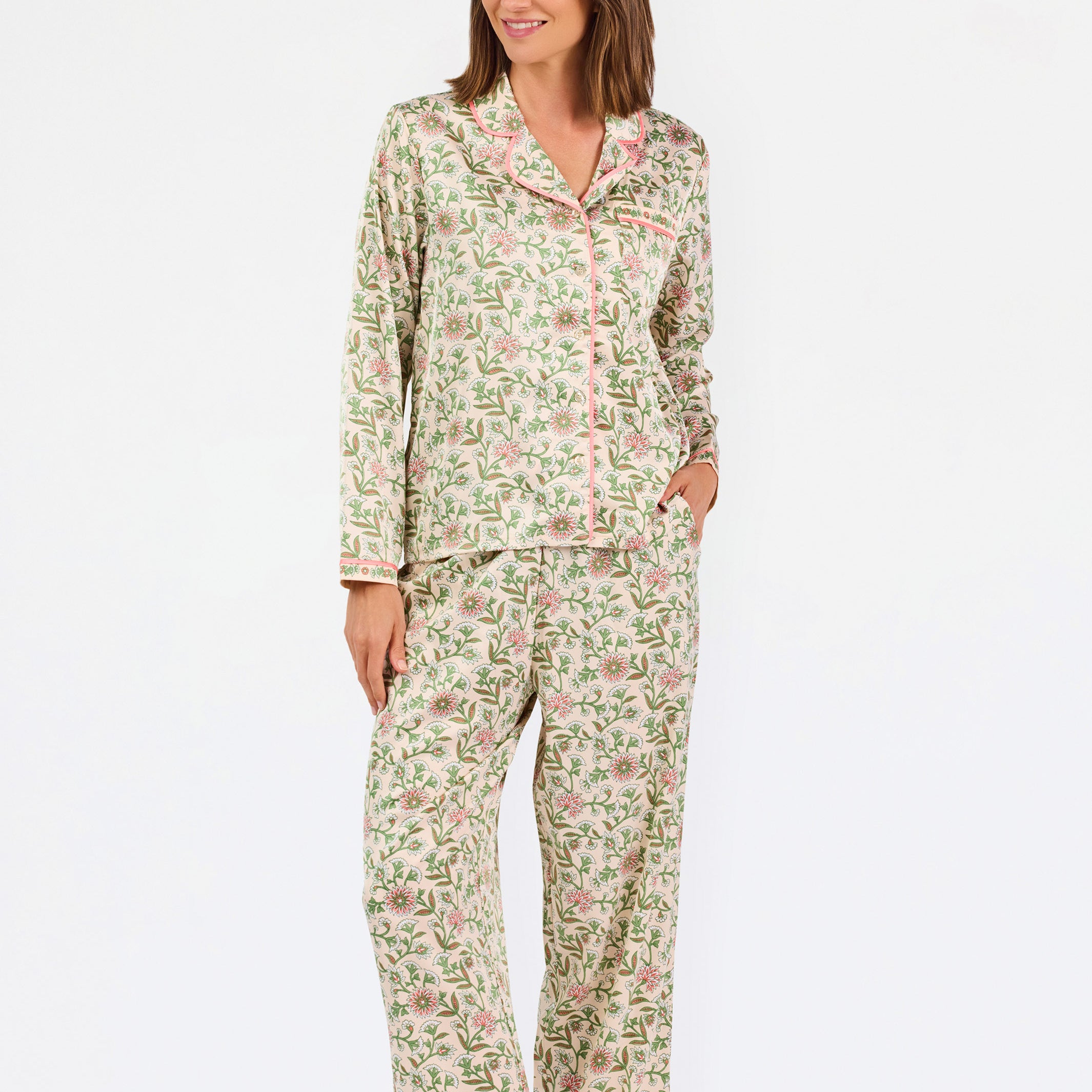 A woman modeling a two-piece pajama set with a long-sleeve button-front top and matching wide-leg pants, featuring a light beige base with a green and pink floral pattern, set against a plain white studio background.