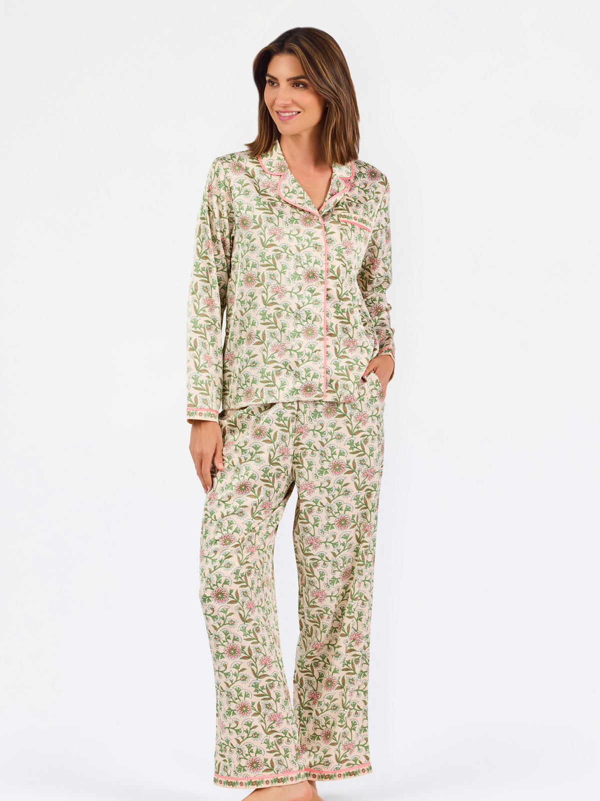 A woman modeling a two-piece pajama set with a long-sleeve button-front top and matching wide-leg pants, featuring a light beige base with a green and pink floral pattern, set against a plain white studio background.