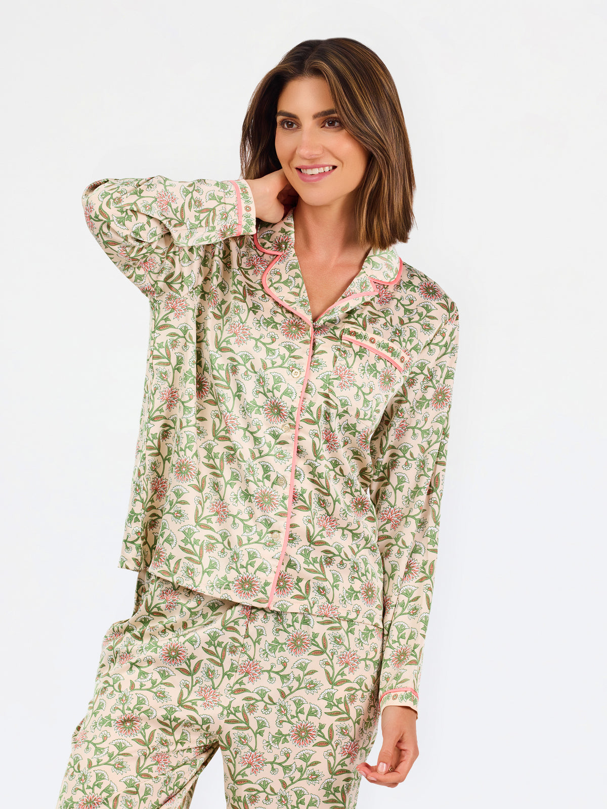 A woman modeling a two-piece pajama set with a long-sleeve button-front top and matching wide-leg pants, featuring a light beige base with a green and pink floral pattern, set against a plain white studio background.
