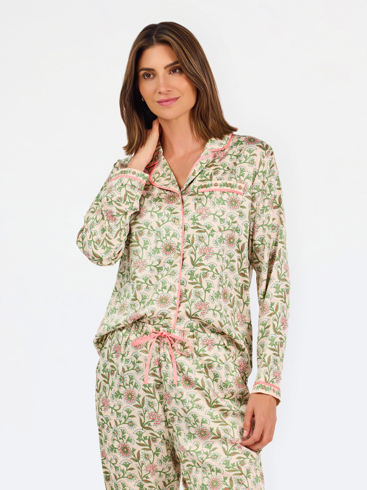 A woman modeling a two-piece pajama set with a long-sleeve button-front top and matching wide-leg pants, featuring a light beige base with a green and pink floral pattern, set against a plain white studio background.
