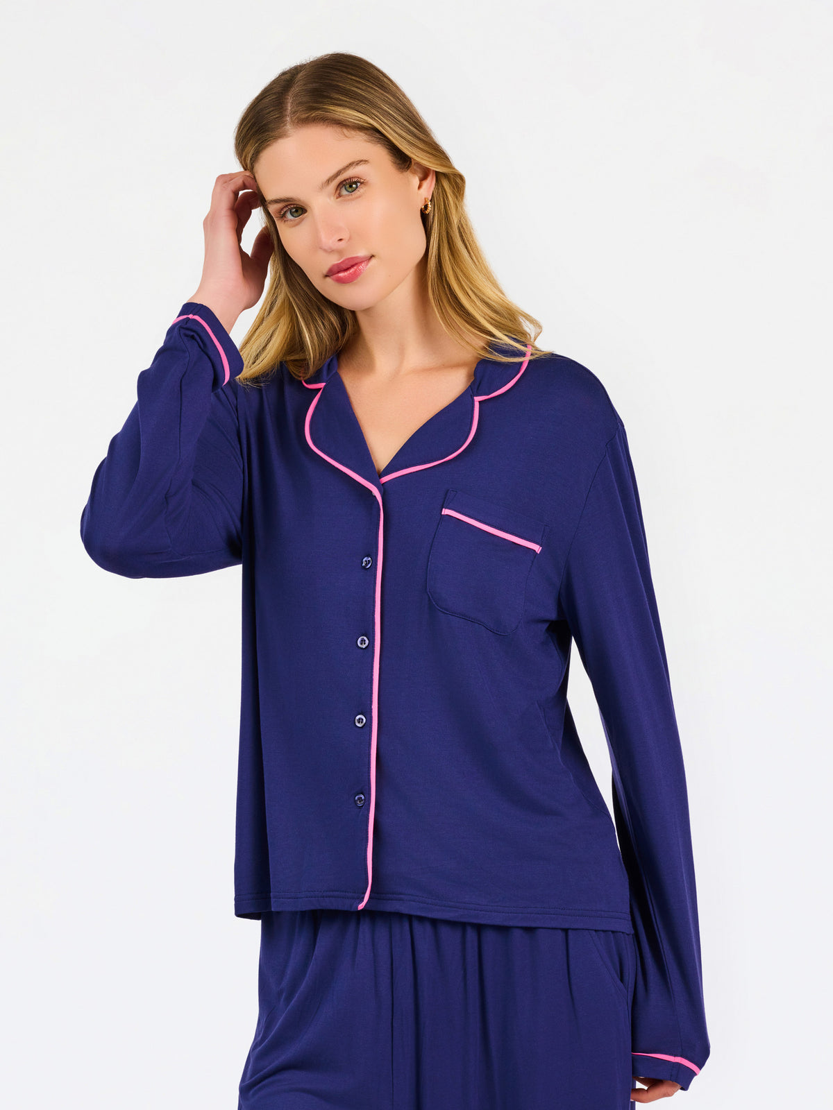 A woman modeling a two-piece pajama set in navy blue with bright pink piping trim, featuring a long-sleeve button-front top and matching pants.