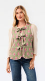 A woman modeling a sleeveless quilted vest in sage green with a pink floral pattern and bow details, worn over a cream puff-sleeve top and blue jeans against a white studio background.