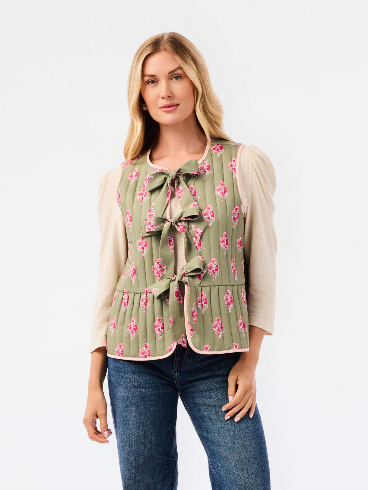 A woman modeling a sleeveless quilted vest in sage green with a pink floral pattern and bow details, worn over a cream puff-sleeve top and blue jeans against a white studio background.