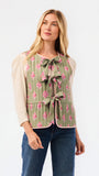 A woman modeling a sage green sleeveless vest with a pink floral pattern, tie-front bow details, and a ruffled hem, worn over a cream puff-sleeve top and blue jeans against a white studio background.