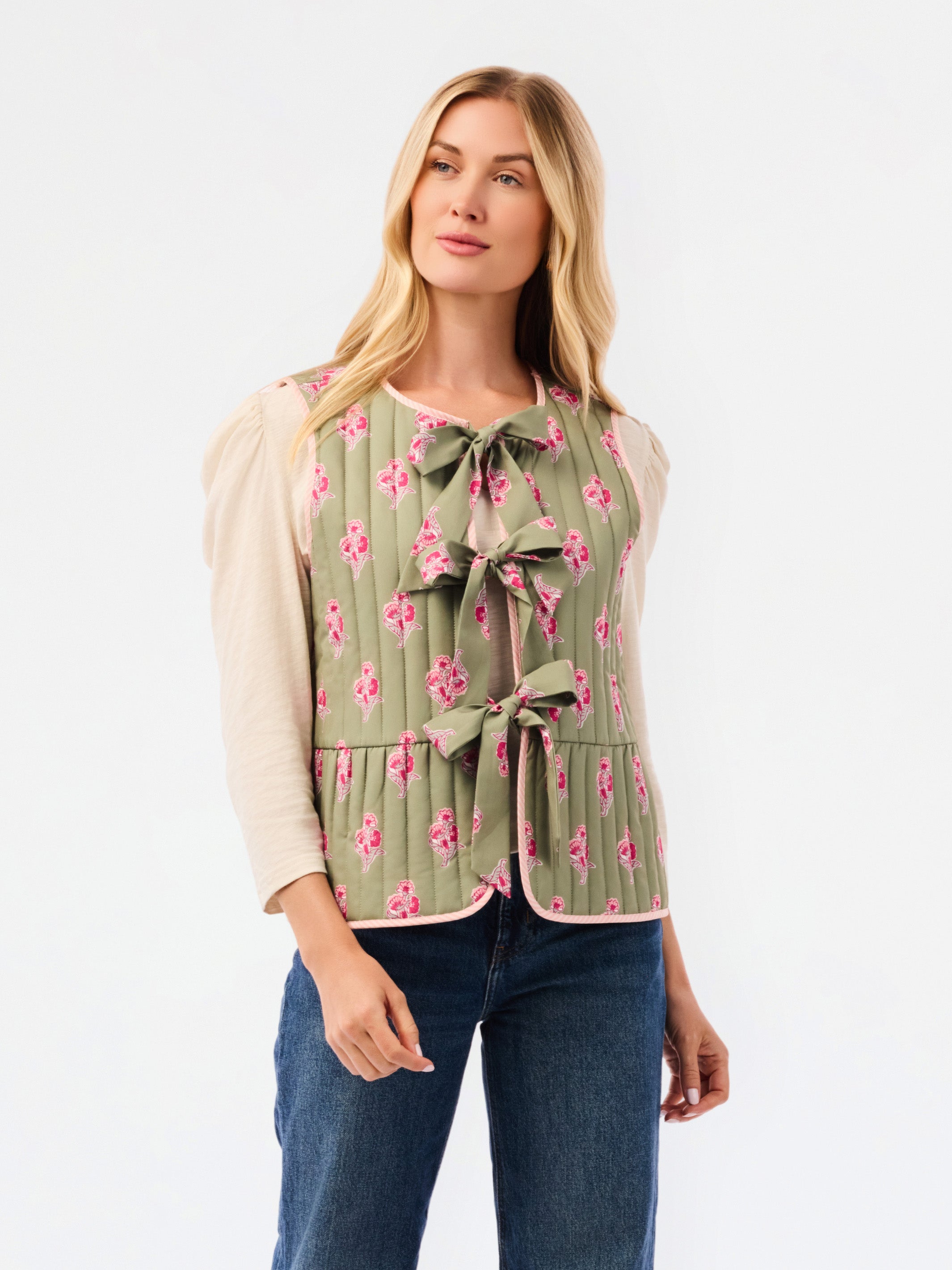 A woman modeling a sage green sleeveless vest with a pink floral pattern, tie-front bow details, and a ruffled hem, worn over a cream puff-sleeve top and blue jeans against a white studio background.