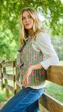 A woman leans against a wooden fence outdoors, wearing a sage green sleeveless vest with a pink floral print over a white long-sleeve shirt and blue jeans.