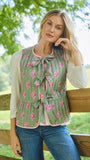 A woman wearing a sage green tiered ruffle vest with a pink floral print, layered over a white long-sleeve shirt and blue jeans, leaning against a wooden fence in an outdoor grassy field.