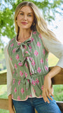 A woman wearing a sage green sleeveless blouse with a pink floral print and a front tie detail, layered over a white long-sleeve top and paired with blue jeans, posing outdoors against a wooden fence with a green, blurred background.