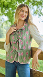 A woman wearing a sage green sleeveless vest with a pink floral print and tie-neck detail, styled over a white long-sleeve shirt and blue jeans, standing outdoors next to a wooden fence.