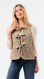 A woman modeling a sleeveless olive green vest with a pink floral print, pleated texture, and front bow details, paired with a cream long-sleeve top and blue jeans against a white studio background.