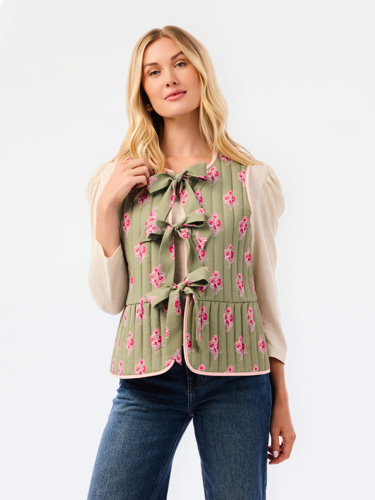 A woman modeling a sleeveless olive green vest with a pink floral print, pleated texture, and front bow details, paired with a cream long-sleeve top and blue jeans against a white studio background.