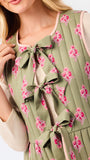 A close-up of a model wearing a sage green quilted vest with pink floral prints and large bow details, layered over a beige long-sleeve top.